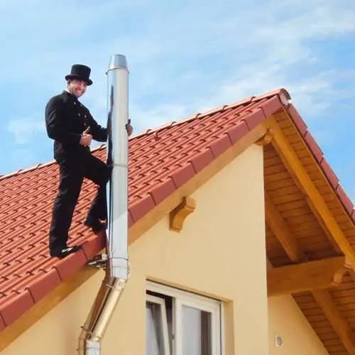 Schornsteinfeger in traditioneller Kleidung steht auf einem Ziegeldach neben einem Edelstahlschornstein.