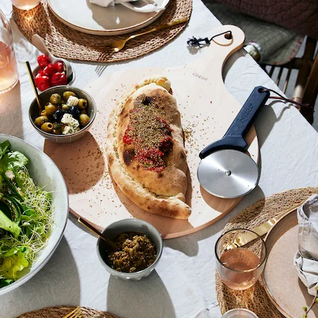 Gedeckter Tisch mit einem belegten Fladenbrot, Oliven, Salat und einem Pizzaschneider Gedeckter Tisch mit einem belegten Fladenbrot, Oliven, Salat und einem Pizzaschneider