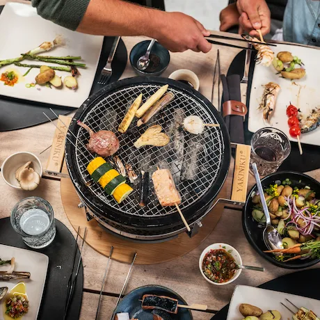 Personen grillen verschiedene Lebensmittel auf einem runden Tischgrill Personen grillen verschiedene Lebensmittel auf einem runden Tischgrill